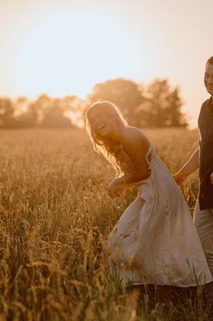 a man and woman holding hands walking through tall grass in front of the setting sun