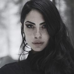 a woman with long black hair and dark makeup looks at the camera while standing in front of trees