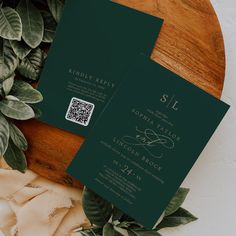 two green wedding cards sitting on top of a wooden table next to some greenery