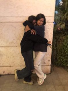 two young women hugging each other on the side of a building with trees in the background