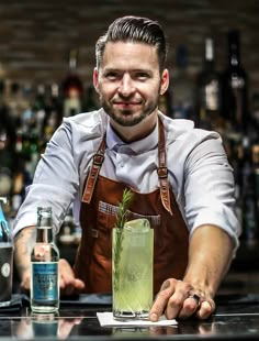 a man in an apron is behind a bar with a green drink and smiling at the camera