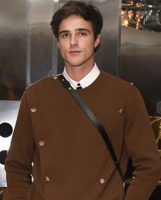 a man standing in front of a metal wall wearing a brown sweater and white shirt