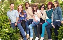 a group of people sitting next to each other on top of a wooden bench surrounded by bushes