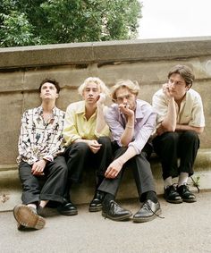 four young men sitting next to each other in front of a stone wall and trees