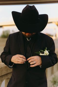 a man in a black suit and cowboy hat is tying his boutonniere