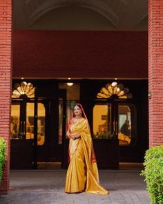 a woman wearing a yellow sari standing in front of a red brick building with the doors open