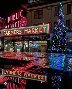 a christmas tree is lit up in front of a farmers market at night with lights reflecting off the wet pavement