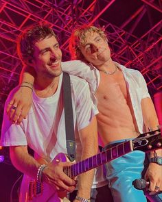 two shirtless men standing next to each other on stage with one holding an electric guitar