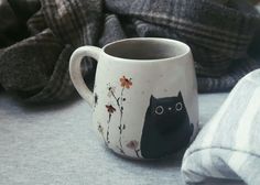 a black cat mug sitting on top of a bed next to a pile of blankets