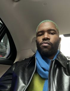 a man with green hair wearing a black jacket and blue scarf in the back seat of a car