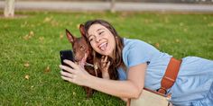 a woman laying on the grass with her dog taking a selfie while holding a cell phone