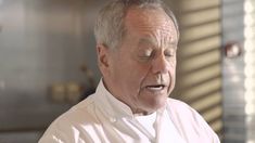 an older man wearing a chef's coat and looking at the camera with his eyes closed