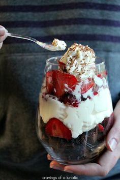 a person is holding a dessert in a glass with strawberries and whipped cream on top