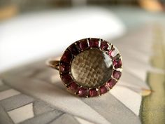 An early nineteenth century memorial ring, set with a cental locket with plaited light brown hair surrounded by a halo of flat cut almandine garnets, set closed-back and foiled, underside with fluted petal design, knife-edge shank in gold, size 4.5, 2.1 grams, circa 1800. During the early nineteenth century it was common for women to adorn themselves with multiple rings of every shape and style, from amatory rings to mourning rings. As such, a rich diversity of rings from this time period can be Antique Ruby Cabochon Ring Gift, Antique Cabochon Ruby Ring Gift, Antique Cabochon Ruby Ring For Gift, Antique Cabochon Ruby Ring For Wedding, Victorian Style Cabochon Ruby Ring Gift, Antique Ruby Ring With Intaglio For Gift, Victorian Round Jewelry With Halo Setting, Antique Jewelry With Halo Setting As Gift, Antique Halo Setting Jewelry As A Gift