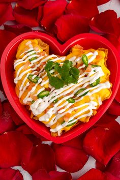 a heart - shaped dish in a red bowl surrounded by flowers and petals, with a green garnish on top