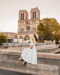 a woman in white dress and hat sitting on the edge of a wall near a cathedral