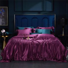 a bed with purple sheets and pillows in a dark colored room next to a blue headboard