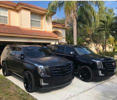 two black suvs parked in front of a house with palm trees and a driveway