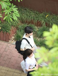 two young women walking down a sidewalk next to trees and bushes, one carrying a black backpack