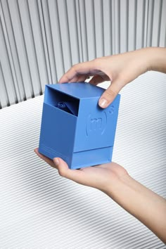 two hands holding a blue box with the letter m in it's middle and one hand on top