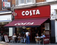 people sitting at tables in front of a restaurant with red awnings on the street