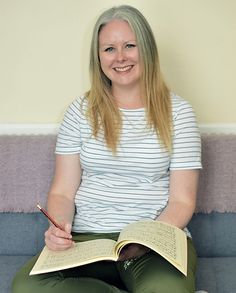 a woman sitting on a couch with a book and pen in her lap while smiling at the camera