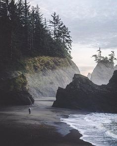 a person walking on the beach next to some rocks and water with trees in the background