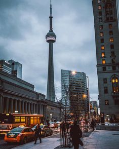 people are walking on the sidewalk in front of some tall buildings at dusk with cars parked nearby