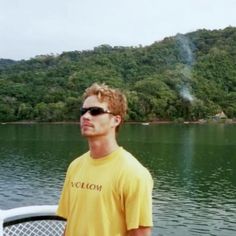 a man standing on top of a boat in front of a body of water with trees behind him