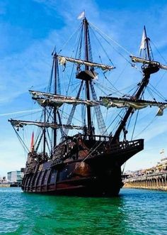 an old pirate ship in the water near a dock with buildings and blue sky behind it