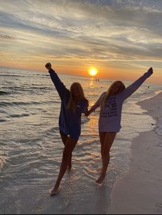 two women are standing on the beach with their arms in the air as the sun sets