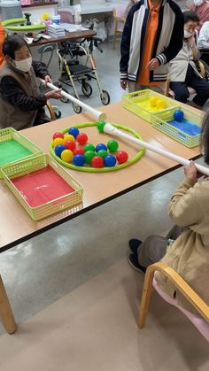 several children sitting at a table with plastic trays and balls on them, while adults watch