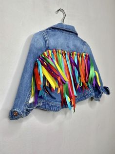 a denim jacket with multicolored fringes hanging on a hook in a white wall