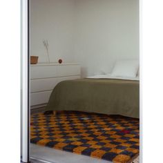 a bed sitting next to a white dresser under a window in a room with an orange and black checkered rug on the floor