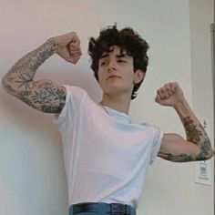 a man with tattoos standing in front of a wall flexing his arms and looking at the camera