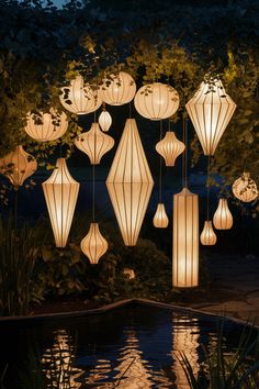 Lanterns of various shapes and sizes illuminated and hanging in a garden at night, reflecting in a pond below. Garden Wedding Lights Outdoor Parties, Whimsical Garden Lighting, Outdoor Floor Lighting Ideas, Lanterns In Garden, Hanging Garden Lights, Tree Lanterns Hanging Backyard, Backyard Lanterns Hanging, Japanese Outdoor Lighting, Garden For Entertaining