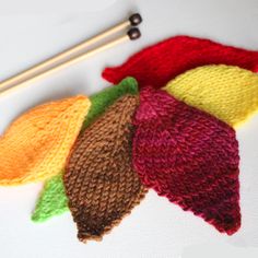 four knitted autumn leaves next to knitting needles and an orange, yellow, green, red, and brown hat