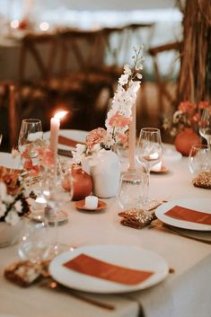 the table is set with white and orange plates, silverware, candles and flower centerpieces