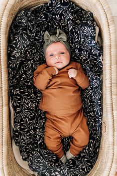 a baby is laying in a basket with a bow on it's head and wearing a brown outfit