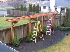 two ladders are next to a wooden arbor in a yard with grass and bushes