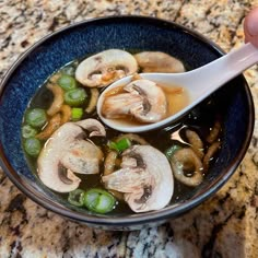 a person holding a spoon over a bowl of soup with mushrooms and broccoli