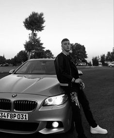 a man sitting on the hood of a silver car in front of a park with trees