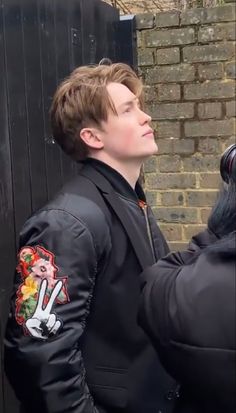 a young man in a black jacket standing next to a brick wall