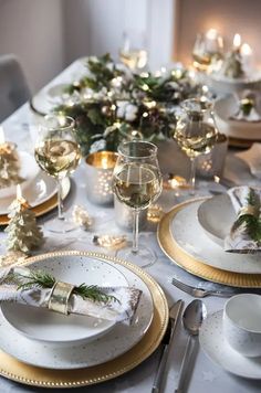 the table is set for christmas dinner with white and gold plates, silverware, candles and greenery