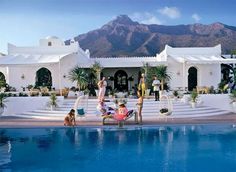 people relaxing by the pool in front of a large white house with mountains in the background