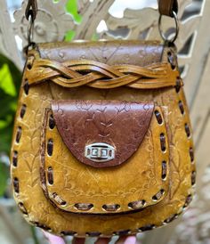 This shoulder bag is hand crafted by an artisan in the 1970s. Features braided flap closure and outside coin purse with metal clasp toggle. Subtle tooling and etching. Two tone golden hue with brown hued leather. Braided strap with o ring fasteners. Long enough for crossbody use on a petite frame. Artisan Hand Tooled Crossbody Saddle Bag, Artisan Handmade Brown Saddle Bag, Handmade Artisan Brown Saddle Bag, Artisan Brown Handmade Saddle Bag, Vintage Brown Crossbody Flap Bag, Artisan Brown Shoulder Bag, Artisan Brown Satchel For Festival, Vintage Hand-stitched Satchel, Brown Artisan Saddle Shoulder Bag