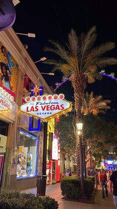 the las vegas sign is lit up at night in front of some shops and palm trees