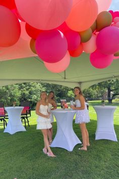 two women in white dresses standing under a tent with balloons hanging from it's ceiling