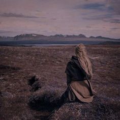 a woman with long hair sitting on top of a grass covered field next to mountains