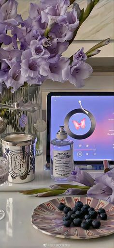 purple flowers are in vases next to a computer screen and blueberries on a plate